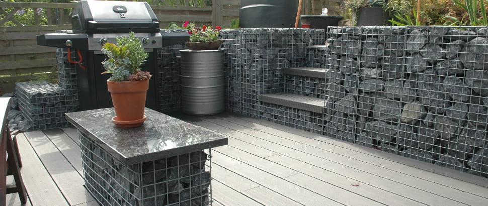 Tuin met schanskorven als trap en muurelementen, gecombineerd met terras en barbecue.