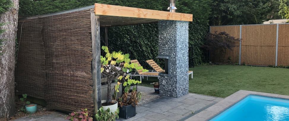 Schanskorfzuilen bieden de perfecte balans tussen design en functionaliteit. Een schanskorfzuil die gebruikt wordt als steunpilaar voor een overkapping in een groene tuin met zwembad.