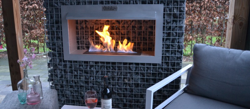 Een stijlvolle bio ethanol haard geïntegreerd in een schanskorf, geplaatst onder een overkapping met gezellige zithoek.