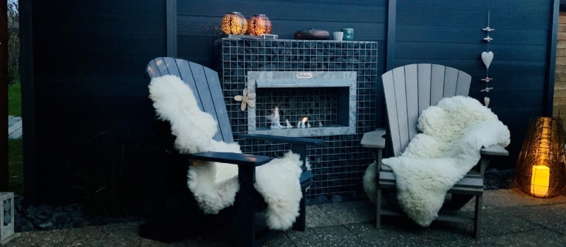 Een sfeervolle bio ethanol haard verwerkt in een schanskorf, geplaatst tussen twee comfortabele stoelen met schapenvacht, in een moderne tuinsetting.