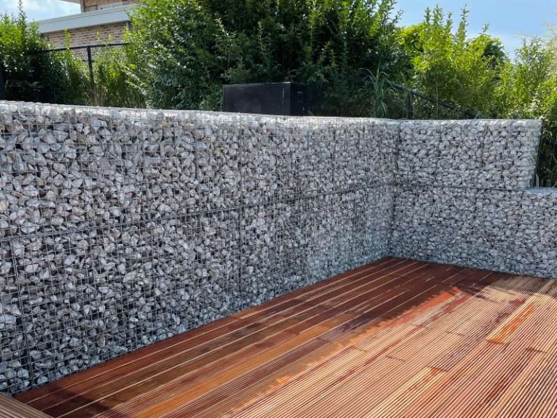 Robuust, stijlvol en tijdloos. Dat is de kracht van schanskorven Houten veranda omgeven met een erfafscheiding bestaande uit schanskorven gevuld met breukstenen